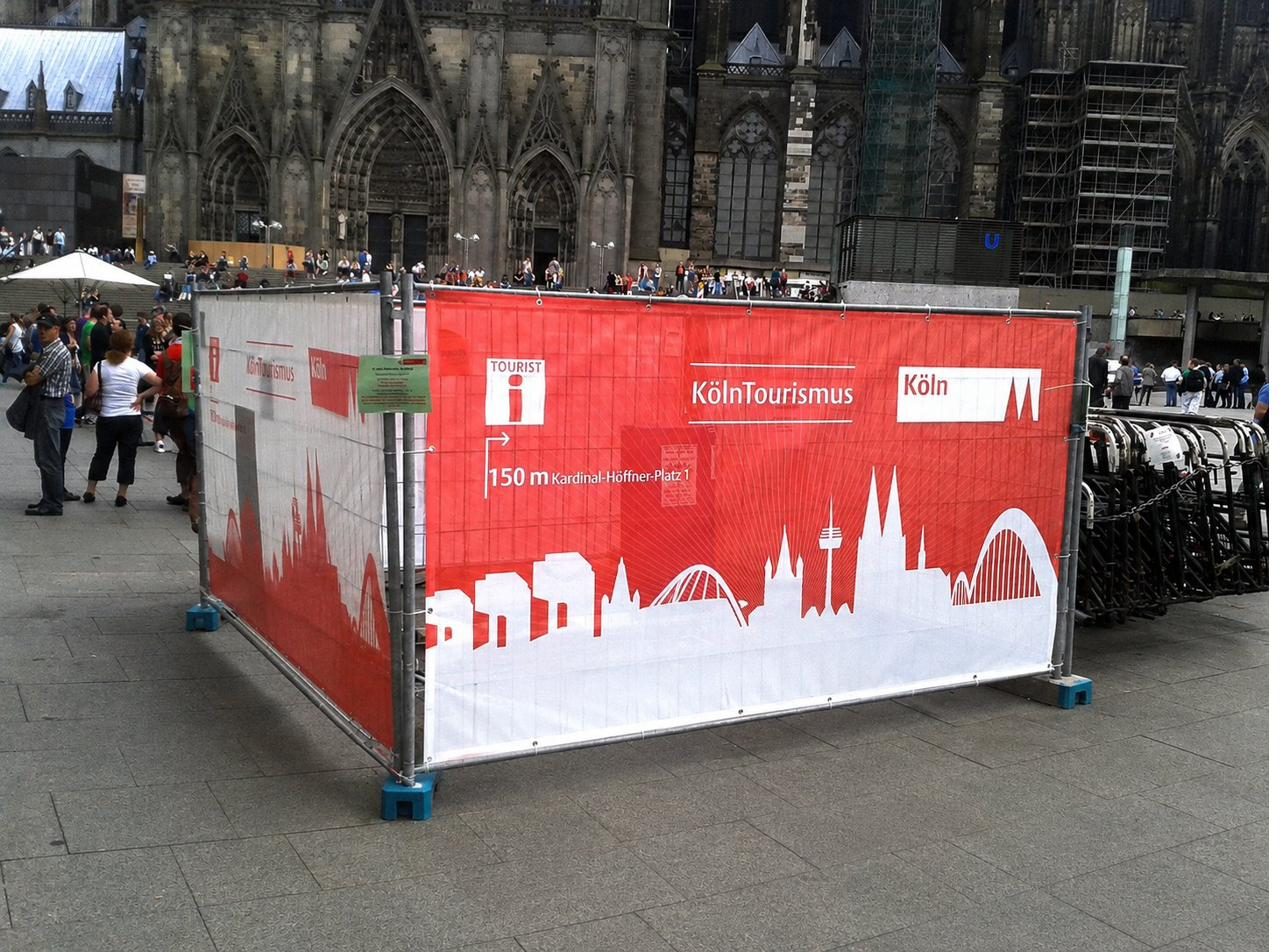 Roter Bauzaunbanner von KölnTourismus mit Kölner Skyline und Entfernungsangabe vor dem Kölner Dom
