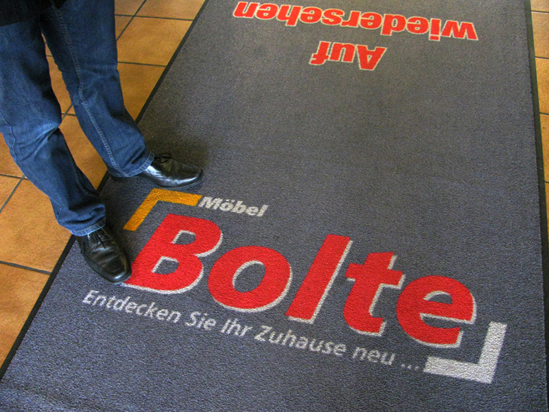 Schmutzfangmatte mit Logo und zweitem Schriftzug in Gegenrichtung, darauf stehende Person